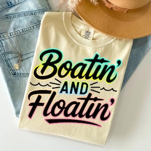K&ouml;nnte beinhalten: Cremefarbenes T-Shirt mit den Worten "Boatin' and Floatin'" in einer farbenfrohen, verspielten Schriftart. Das Shirt ist gefaltet und liegt auf einer blauen Jeans, einer Halskette und einem braunen Hut. Das Shirt ist aus weichem Material.