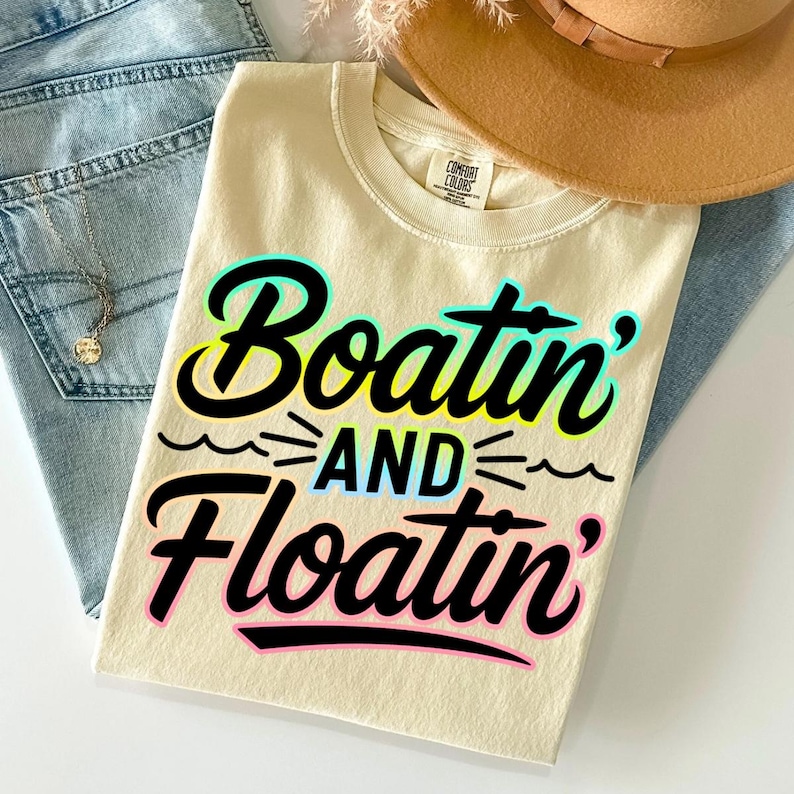 K&ouml;nnte beinhalten: Cremefarbenes T-Shirt mit den Worten "Boatin' and Floatin'" in einer farbenfrohen, verspielten Schriftart. Das Shirt ist gefaltet und liegt auf einer blauen Jeans, einer Halskette und einem braunen Hut. Das Shirt ist aus weichem Material.