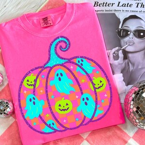 May include: A bright pink t-shirt featuring a Halloween-themed design. The graphic includes a large pumpkin shape with teal ghosts and jack-o'-lanterns. The pumpkin is outlined in purple glitter, with a teal stem. The shirt is made of a soft material.