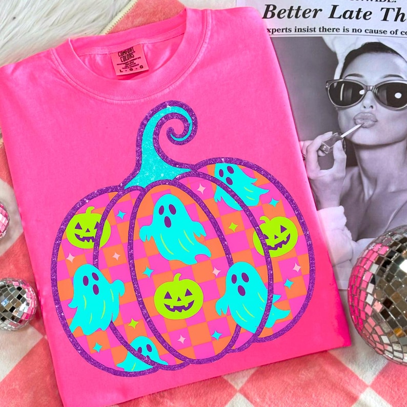 May include: A bright pink t-shirt featuring a Halloween-themed design. The graphic includes a large pumpkin shape with teal ghosts and jack-o'-lanterns. The pumpkin is outlined in purple glitter, with a teal stem. The shirt is made of a soft material.