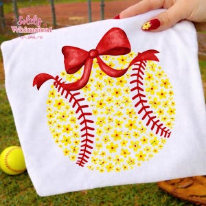 May include: White t-shirt featuring a softball design. The softball is filled with yellow flowers and red stitching, topped with a red bow. A yellow softball is visible in the lower left corner. The text "Solely Whimsical" is in the upper left corner.