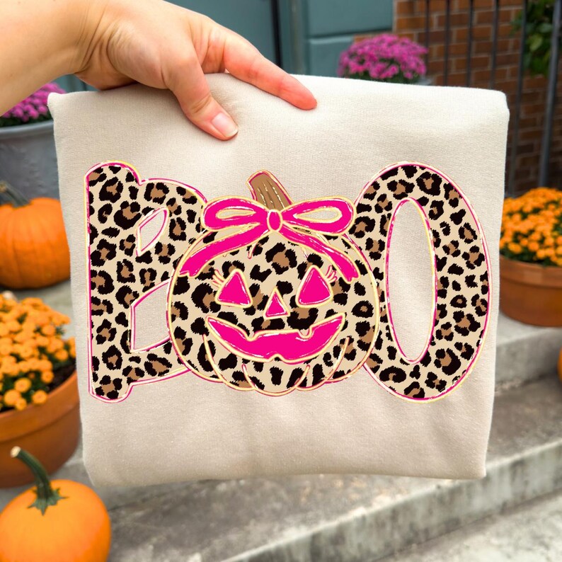 Op de afbeelding: Een beige sweatshirt met een Halloween-thema. Het woord "BOO" is in luipaardprint, met een roze omrande pompoen in het midden. De pompoen heeft een roze strik en een lachend gezicht.