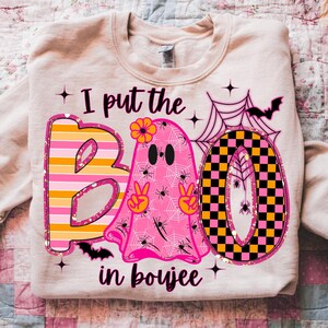 May include: A light beige sweatshirt with a Halloween-themed graphic. The graphic includes the words "I put the BOO in boujee" with a pink ghost, spiderwebs, bats, and the letters B and O in a colorful design.