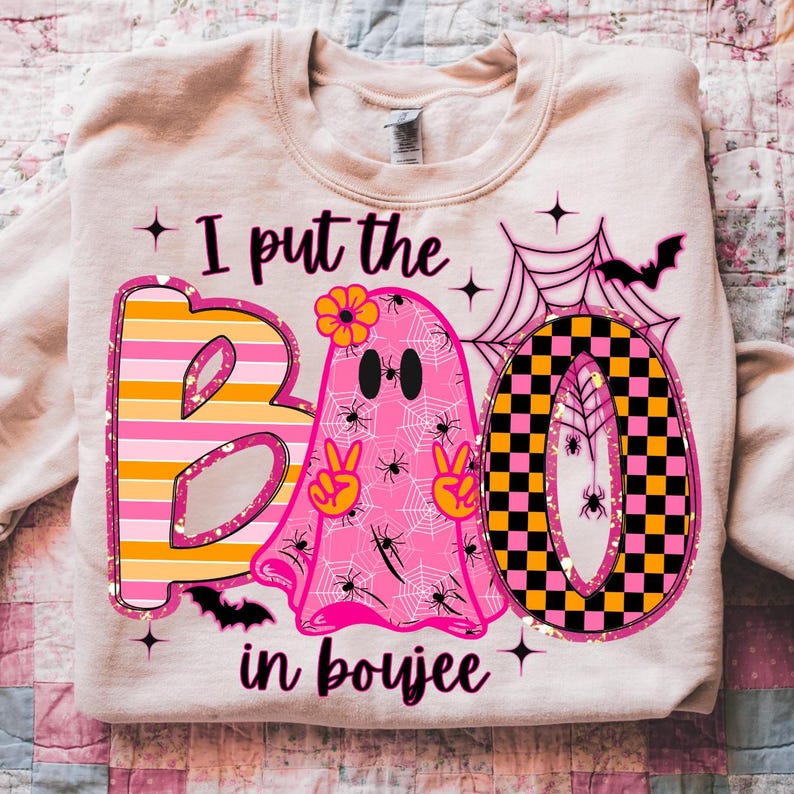 May include: A light beige sweatshirt with a Halloween-themed graphic. The graphic includes the words "I put the BOO in boujee" with a pink ghost, spiderwebs, bats, and the letters B and O in a colorful design.