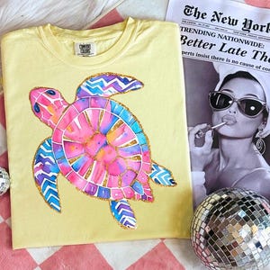 May include: Yellow Comfort Colors t-shirt featuring a colorful watercolor sea turtle design. The turtle has pink, blue, and gold accents. The shirt is displayed with a newspaper and a disco ball.