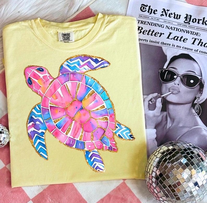 May include: Yellow Comfort Colors t-shirt featuring a colorful watercolor sea turtle design. The turtle has pink, blue, and gold accents. The shirt is displayed with a newspaper and a disco ball.