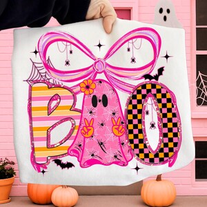 Pode incluir: Cobertor branco com tema de Halloween com a palavra "BOO" em um design l&uacute;dico. O "B" &eacute; listrado, o "O" tem um padr&atilde;o xadrez e o segundo "O" &eacute; um fantasma rosa com sinais de paz. O design inclui aranhas, morcegos e um grande la&ccedil;o.