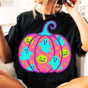 May include: Black t-shirt featuring a large, colorful Halloween pumpkin design. The pumpkin has a purple outline, teal stem, and a checkered orange and pink pattern. Teal ghosts and green jack-o'-lanterns are also featured.