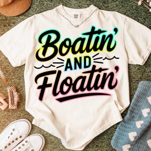 K&ouml;nnte beinhalten: Cremefarbenes T-Shirt mit dem Schriftzug "Boatin' and Floatin'" in einer farbenfrohen, verspielten Schriftart. Der Text ist schwarz umrandet, mit einem t&uuml;rkis-gelben Farbverlauf. Das Shirt ist Teil einer Casual-Wear-Kollektion.