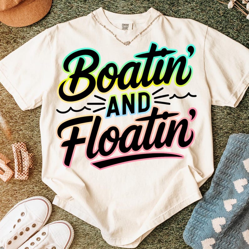 K&ouml;nnte beinhalten: Cremefarbenes T-Shirt mit dem Schriftzug "Boatin' and Floatin'" in einer farbenfrohen, verspielten Schriftart. Der Text ist schwarz umrandet, mit einem t&uuml;rkis-gelben Farbverlauf. Das Shirt ist Teil einer Casual-Wear-Kollektion.