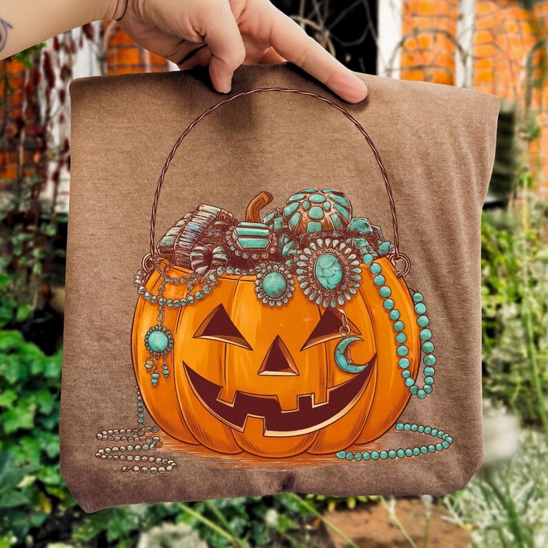 May include: Brown t-shirt featuring a smiling jack-o'-lantern with turquoise jewelry and a crescent moon. The pumpkin has a brown handle and is filled with necklaces and bracelets. The design is a Halloween theme.