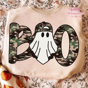 以下が含まれることがあります： ハロウィーンをテーマにしたデザインのベージュのスウェットシャツ。「BOO」の文字は迷彩柄で、真ん中に迷彩柄の野球帽をかぶった幽霊がいます。幽霊は黒い目と白い体を持っています。