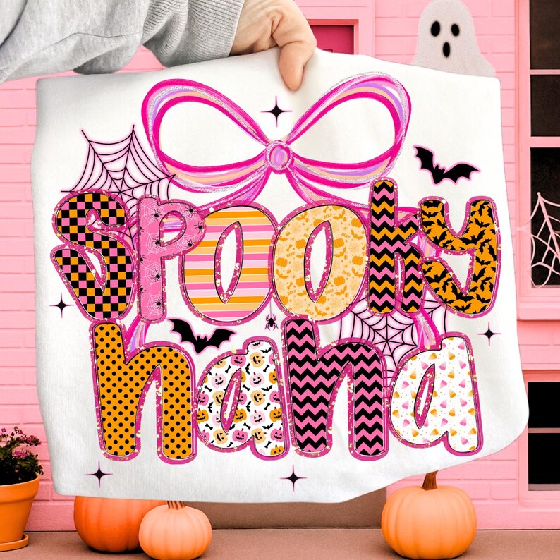Peut inclure: Une couverture blanche sur le th&egrave;me d'Halloween avec les mots "Spooky Nana" dans un motif color&eacute;. Le motif comprend un n&oelig;ud rose, des toiles d'araign&eacute;es, des chauves-souris et des &eacute;toiles. Deux citrouilles sont devant la couverture.