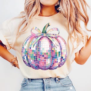 Puede incluir: Camiseta color crema con un dise&ntilde;o de calabaza de bola de discoteca colorida. La calabaza est&aacute; formada por peque&ntilde;as baldosas cuadradas en tonos rosas, morados, azules y verdes, con un tallo verde y una cinta pastel. La camiseta es de estilo casual.