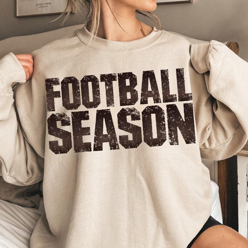 May include: A beige sweatshirt featuring the text "FOOTBALL SEASON" in a distressed, dark brown font. The sweatshirt has a crew neck and long sleeves. The style is casual and comfortable, suitable for cooler weather.