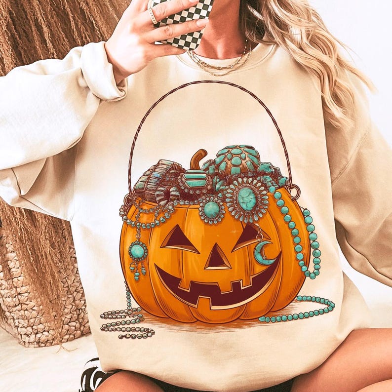 May include: Beige sweatshirt featuring a jack-o'-lantern design filled with turquoise jewelry. The pumpkin has a carved face and is adorned with necklaces and bracelets. A black handle arches over the pumpkin.