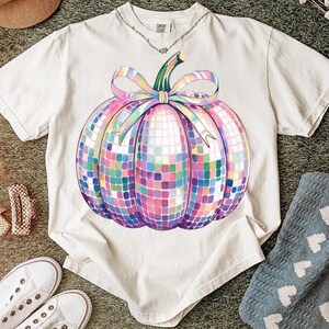 Puede incluir: Camiseta color crema con un dise&ntilde;o de calabaza de bola de discoteca colorida. La calabaza tiene un tallo verde y un gran lazo de colores pastel. La camiseta est&aacute; hecha de un material suave y es adecuada para el uso diario.