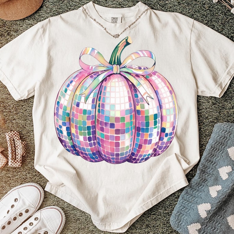 Puede incluir: Camiseta color crema con un dise&ntilde;o de calabaza de bola de discoteca colorida. La calabaza tiene un tallo verde y un gran lazo de colores pastel. La camiseta est&aacute; hecha de un material suave y es adecuada para el uso diario.