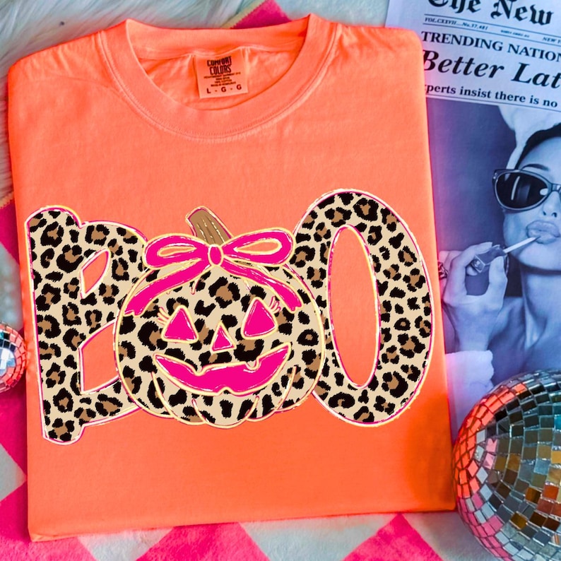 Op de afbeelding: Fel oranje t-shirt met een luipaardprint "BOO" ontwerp en een roze pompoen. De pompoen heeft een roze strik en een lachend gezicht. Een leuke Halloween-thema grafische tee.