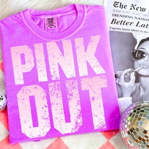 Puede incluir: Una camiseta rosa brillante con las palabras "PINK OUT" en una fuente rosa claro desgastada. La camiseta est&aacute; doblada y colocada sobre una superficie a cuadros rojos y blancos. Un recorte de peri&oacute;dico y una bola de discoteca est&aacute;n en el fondo.