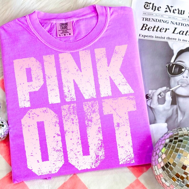 Puede incluir: Una camiseta rosa brillante con las palabras "PINK OUT" en una fuente rosa claro desgastada. La camiseta est&aacute; doblada y colocada sobre una superficie a cuadros rojos y blancos. Un recorte de peri&oacute;dico y una bola de discoteca est&aacute;n en el fondo.