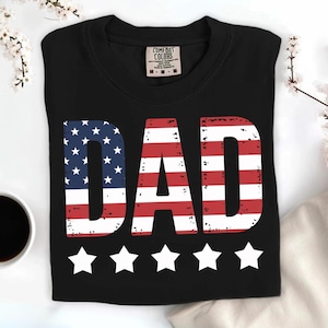 Puede incluir: Camiseta negra con la palabra "DAD" en rojo y blanco, con el dise&ntilde;o de la bandera americana en la "D" y la "A". Cinco estrellas blancas est&aacute;n debajo de la palabra. La camiseta est&aacute; doblada y tiene una etiqueta Comfort Colors.