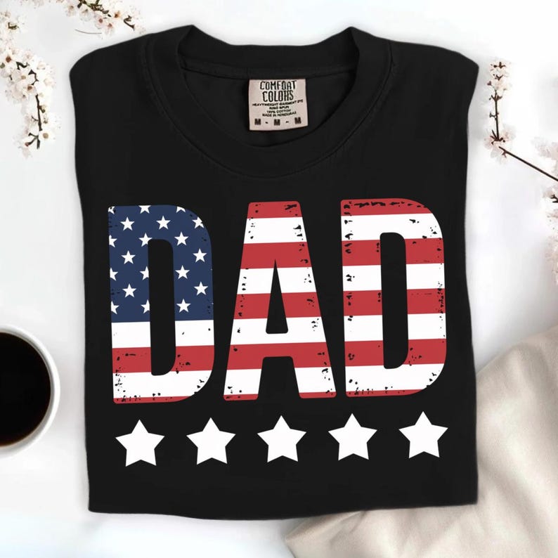 Puede incluir: Camiseta negra con la palabra "DAD" en rojo y blanco, con el dise&ntilde;o de la bandera americana en la "D" y la "A". Cinco estrellas blancas est&aacute;n debajo de la palabra. La camiseta est&aacute; doblada y tiene una etiqueta Comfort Colors.