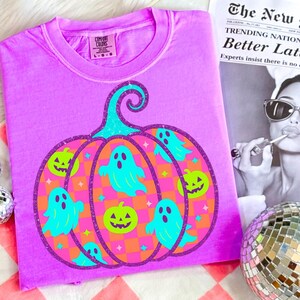 May include: A lilac-colored t-shirt featuring a large pumpkin design with a teal stem. The pumpkin is decorated with teal ghosts, green jack-o'-lanterns, and orange and pink checkered patterns. The shirt has a crew neck and short sleeves.