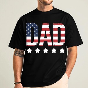 Puede incluir: Camiseta negra con la palabra "DAD" en letras grandes y desgastadas. Las letras est&aacute;n dise&ntilde;adas con el patr&oacute;n de la bandera estadounidense. Cinco estrellas blancas est&aacute;n debajo.