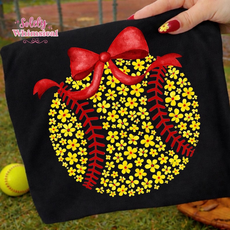 May include: Black t-shirt featuring a baseball design made of yellow flowers and red stitching. A red bow is at the top of the design. A softball is in the background. The text "Solely Whimsical" is in the upper left corner.