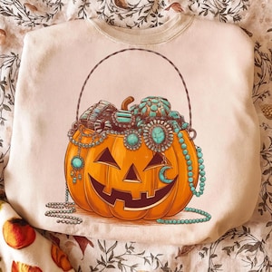 May include: Cream-colored sweatshirt featuring a jack-o'-lantern design. The orange pumpkin is filled with turquoise jewelry and has a smiling face. A crescent moon and beaded necklaces are also included in the design.