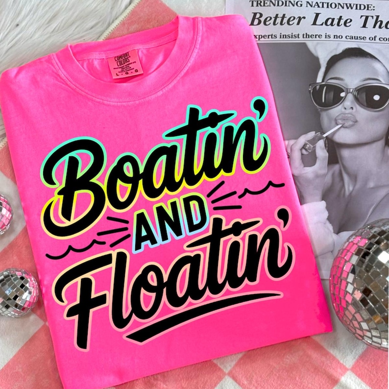 K&ouml;nnte beinhalten: Ein leuchtend pinkfarbenes Sweatshirt mit dem Schriftzug "Boatin' AND Floatin'" in Schwarz, T&uuml;rkis und Gelb. Das Sweatshirt wird auf einer rosa-wei&szlig; karierten Oberfl&auml;che pr&auml;sentiert, mit Discokugeln und einer Zeitung im Hintergrund.