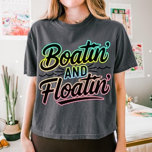 K&ouml;nnte beinhalten: Dunkelgraues T-Shirt mit dem Schriftzug "Boatin' and Floatin'" in einem farbenfrohen Retro-Schriftstil. Der Text ist schwarz umrandet und mit Neonfarben gef&uuml;llt, was ein Sommer- oder Urlaubsthema hervorruft. Das T-Shirt hat eine lockere Passform.