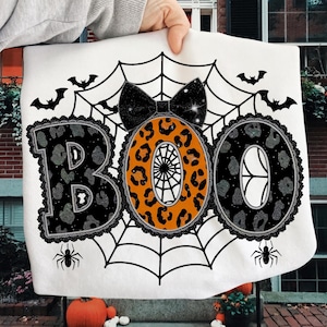 May include: White Halloween-themed sweatshirt with the word "BOO" in a spiderweb design. The letters are decorated with leopard print, a black bow, bats, and spiders. The sweatshirt is held up by a person's hand.