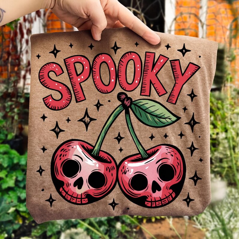 Może przedstawiać: Brązowa torba z upiornym wzorem. Słowo "SPOOKY" jest wydrukowane na czerwono, nad parą wiśni z twarzami czaszek. Czarne gwiazdki otaczają wiśnie i tekst, tworząc motyw Halloween.
