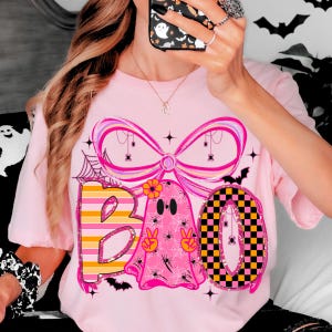 Op de afbeelding: Roze T-shirt met een Halloween-thema. Het woord "BOO" wordt weergegeven met een gestreepte "B", een spook met vredestekens en een geblokte "O". Het ontwerp bevat spinnen, spinnenwebben en een grote roze strik.