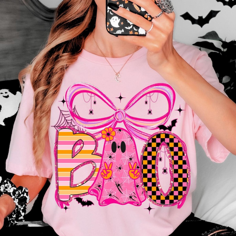 Op de afbeelding: Roze T-shirt met een Halloween-thema. Het woord "BOO" wordt weergegeven met een gestreepte "B", een spook met vredestekens en een geblokte "O". Het ontwerp bevat spinnen, spinnenwebben en een grote roze strik.
