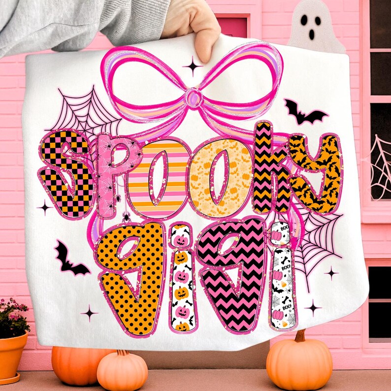 Peut inclure: Une couverture blanche sur le th&egrave;me d'Halloween avec les mots "Spooky Vibes" en rose, orange, noir et jaune. Le motif comprend des chauves-souris, des toiles d'araign&eacute;es et un grand n&oelig;ud rose. La couverture est tenue par une main.