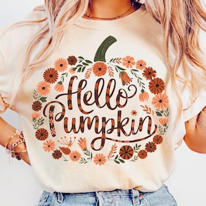 May include: Cream-colored t-shirt featuring the phrase "Hello Pumpkin" in a decorative script, surrounded by a floral wreath in shades of orange, brown, and green. The design is centered on the front of the shirt, creating a seasonal autumn theme.