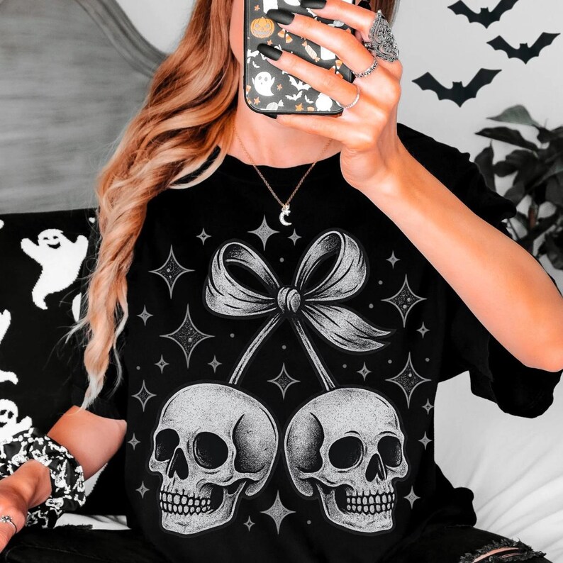 K&ouml;nnte beinhalten: Schwarzes T-Shirt mit einer detaillierten Grafik von zwei Sch&auml;deln, die durch eine Schleife verbunden sind, mit Stern- und Diamantakzenten in Grau und Wei&szlig;. Das Design ist Teil einer Halloween- oder Gothic-Themenkollektion.