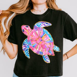 K&ouml;nnte beinhalten: Schwarzes T-Shirt mit einem farbenfrohen Aquarell-Design einer Meeresschildkr&ouml;te. Die Schildkr&ouml;te hat einen rosa und blauen Panzer mit goldenen Akzenten und blau-violetten Flossen. Das Shirt ist ein l&auml;ssiger Stil.