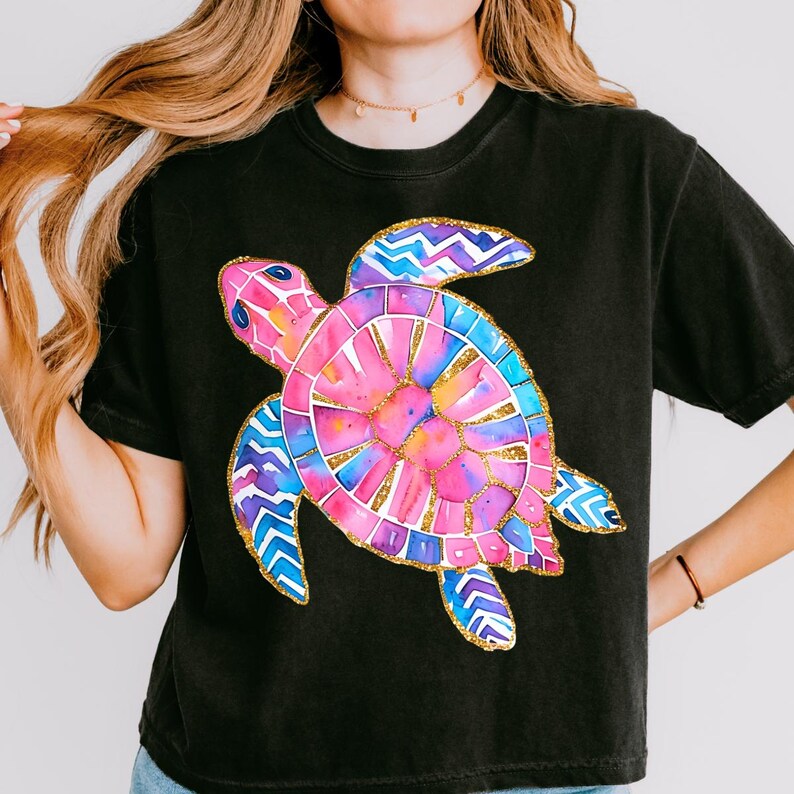K&ouml;nnte beinhalten: Schwarzes T-Shirt mit einem farbenfrohen Aquarell-Design einer Meeresschildkr&ouml;te. Die Schildkr&ouml;te hat einen rosa und blauen Panzer mit goldenen Akzenten und blau-violetten Flossen. Das Shirt ist ein l&auml;ssiger Stil.