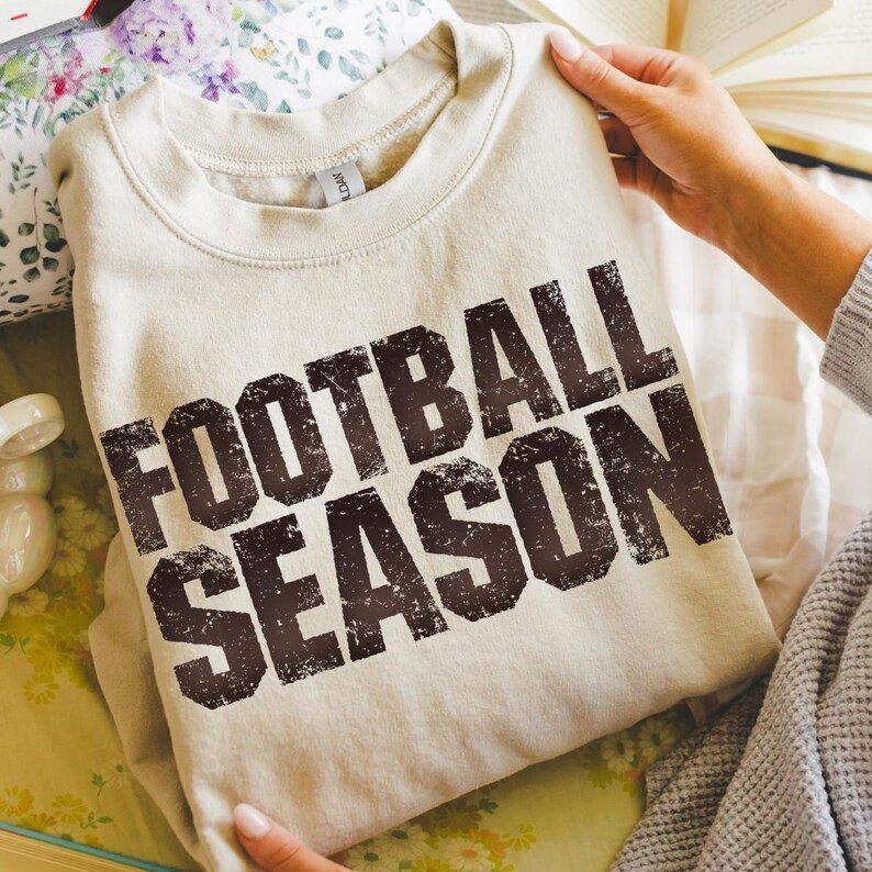 May include: A cream-coloured sweatshirt featuring the text "FOOTBALL SEASON" in distressed brown lettering. The garment is folded and presented on a floral patterned surface.