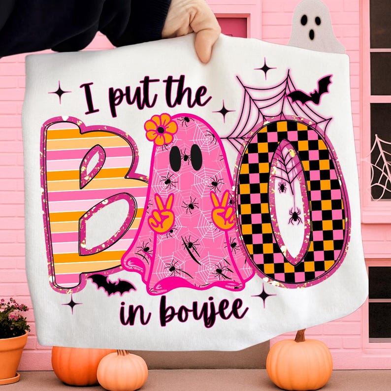 Pode incluir: Cobertor branco com tema de Halloween com a frase "I put the BOO in boujee". O design apresenta um fantasma rosa com sinais de paz, aranhas e uma flor, juntamente com um "O" xadrez preto e amarelo.