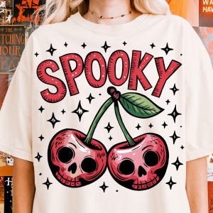 Może przedstawiać: Kremowa koszulka z czerwonym napisem "SPOOKY" i dwiema wiśniami z czaszkami. Zielone liście i łodygi łączą wiśnie. Czarne gwiazdki otaczają tekst i wiśnie.