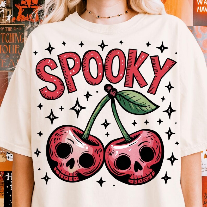 Może przedstawiać: Kremowa koszulka z czerwonym napisem "SPOOKY" i dwiema wiśniami z czaszkami. Zielone liście i łodygi łączą wiśnie. Czarne gwiazdki otaczają tekst i wiśnie.