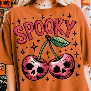 Może przedstawiać: Pomarańczowa koszulka z r&oacute;żową grafiką "SPOOKY" nad dwiema wiśniami z twarzami czaszek. Projekt zawiera zielone łodygi, zielony liść i czarne gwiazdki. Motyw Halloween.