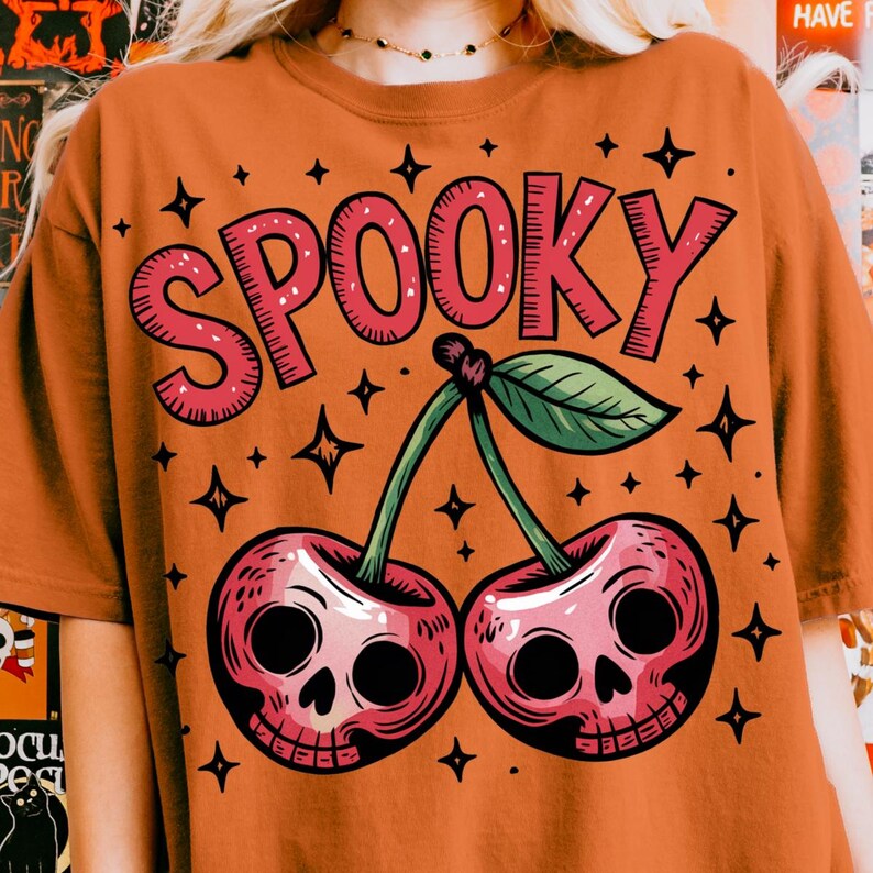 Może przedstawiać: Pomarańczowa koszulka z r&oacute;żową grafiką "SPOOKY" nad dwiema wiśniami z twarzami czaszek. Projekt zawiera zielone łodygi, zielony liść i czarne gwiazdki. Motyw Halloween.