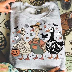 May include: A light gray sweatshirt featuring three cartoon geese in Halloween costumes. One is wrapped in bandages, another wears overalls and a straw hat, and the third is a skeleton. Bats, ghosts, and leaves decorate the design.