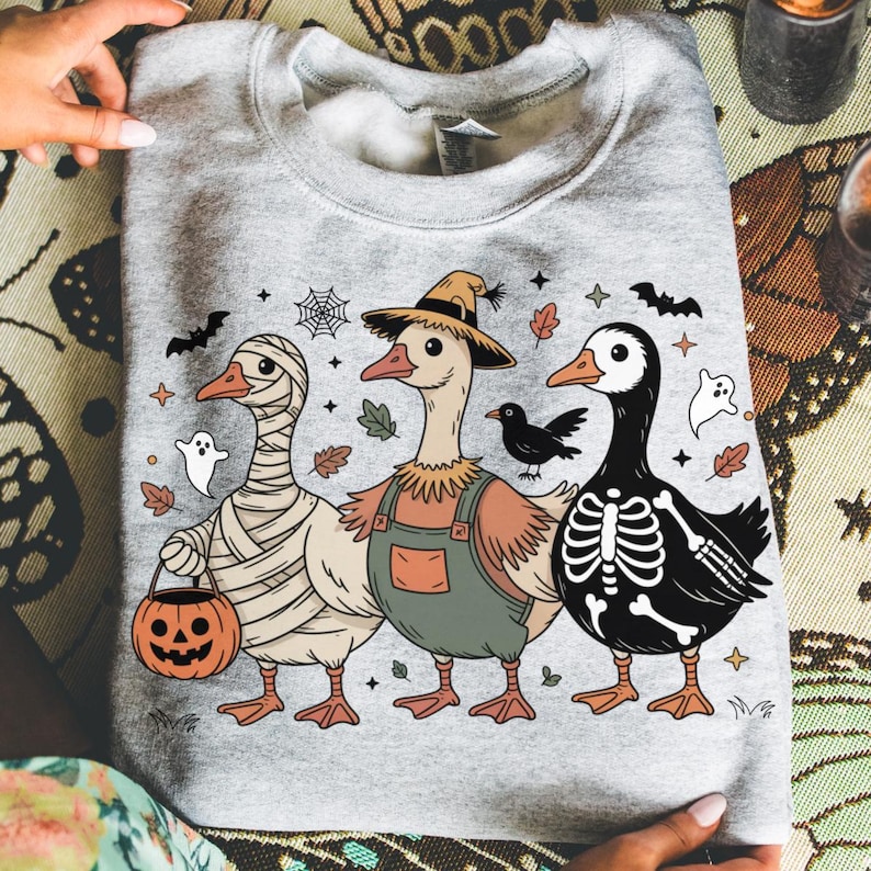 May include: A light gray sweatshirt featuring three cartoon geese in Halloween costumes. One is wrapped in bandages, another wears overalls and a straw hat, and the third is a skeleton. Bats, ghosts, and leaves decorate the design.